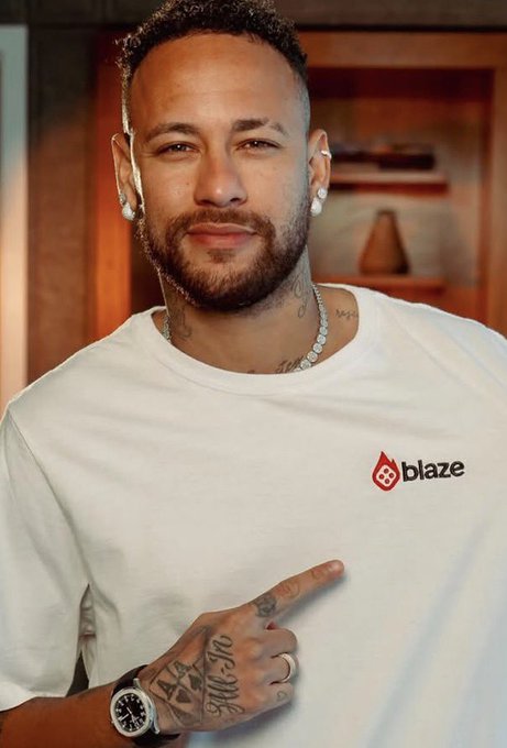 Brazilian soccer player Neymar smiles and points to a Blaze logo on his white t-shirt while wearing a luxury watch and diamond jewelry.