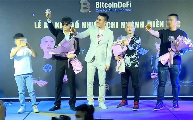 A group of men stand on stage at a 'BitcoinDeFi' event (banner reads 'One for All, All for One'), holding flowers and toasting with glasses in a celebratory moment.
