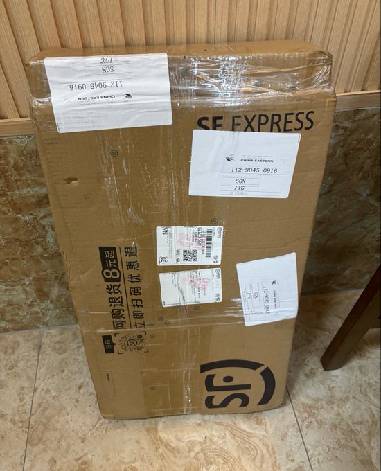 A large cardboard shipping box from SF Express with shipping labels reading 'CHINA EASTERN', 'SGN', and 'PVG' and a phone number, sitting on a tiled floor.