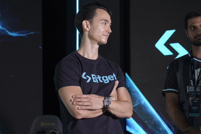 A man wearing a black 'Bitget' t-shirt with crossed arms stands smiling, with another person in a black jersey visible to the right against a dark stage background.