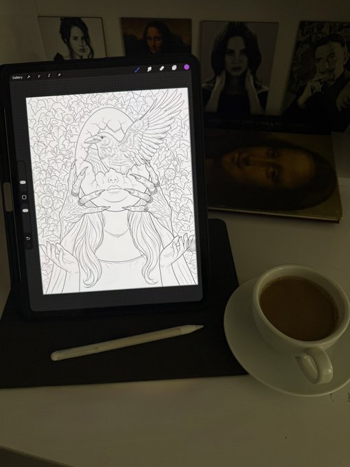 An iPad displaying a digital line art drawing of a girl with a bird on her hands, sitting on a desk next to a stylus, a cup of coffee, and portrait photographs.