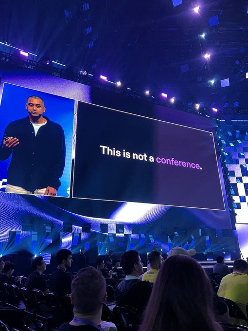A speaker on stage at a large conference with a giant screen displaying 'This is not a conference,' while an audience watches from their seats in a darkened auditorium.