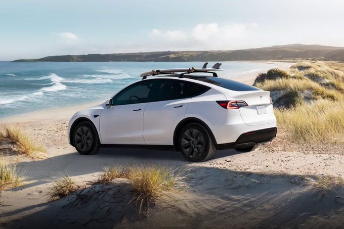A white Tesla Model Y with a roof rack and surfboard is parked on a sandy beach with dunes and the ocean in the background.