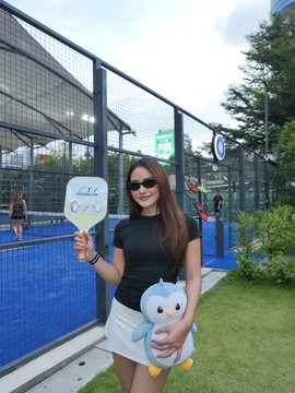 A photo of a woman in a black top and white skirt holding a paddle sign and a stuffed penguin toy at what appears to be a padel or tennis court.