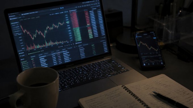 A dimly lit workspace features a laptop displaying a financial trading chart with red and green candlesticks, accompanied by a smartphone showing a similar stock graph, a coffee mug, and a handwritten notebook.