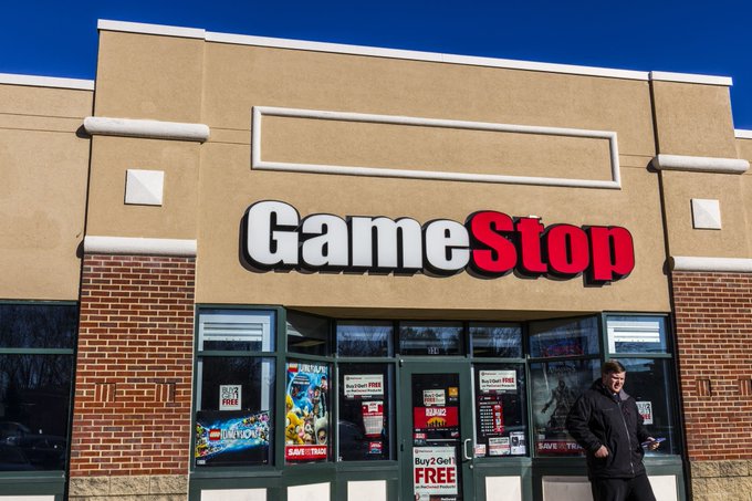 A man walks past a GameStop storefront featuring the company's red and white logo above glass doors displaying promotional posters for video game trade-in deals and "Buy 2 Get 1 FREE" offers, visually reinforcing the financial context of GameStop's substantial $4.95 billion eBay holdings and its expanding digital asset initiatives.
