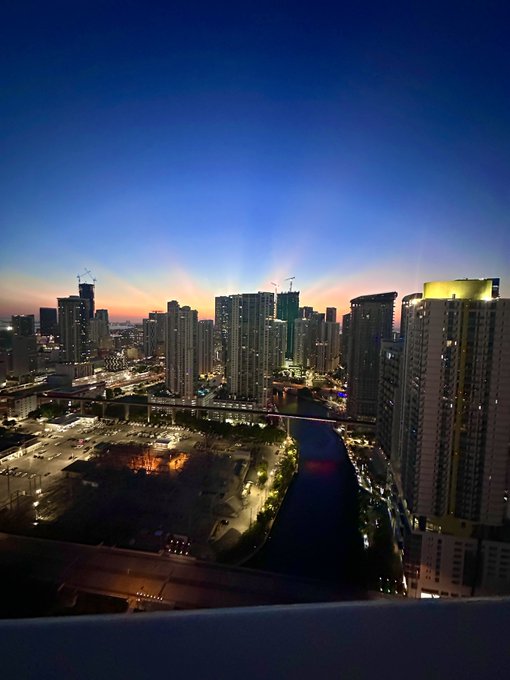 An evening cityscape of high-rise buildings at dusk with a river running through the urban landscape, bridges connecting the city, and the sky transitioning from orange to deep blue.