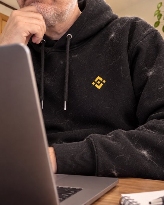 A person in a dusty black hoodie with a small yellow Binance logo sits thoughtfully at a laptop, with visible pet hair on the clothing hinting at a casual, home-office setup. The "IYKYK" caption cleverly nods to Binance users who recognize the subtle brand emblem—a wink to those "in the know" within the crypto community.