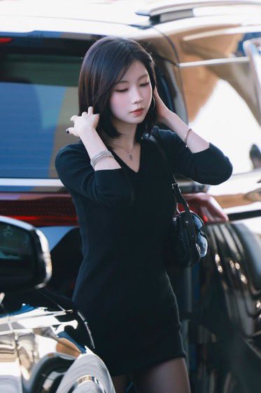 A young woman with dark shoulder-length hair stands beside a car, wearing a fitted black dress and carrying a black handbag with a heart charm.