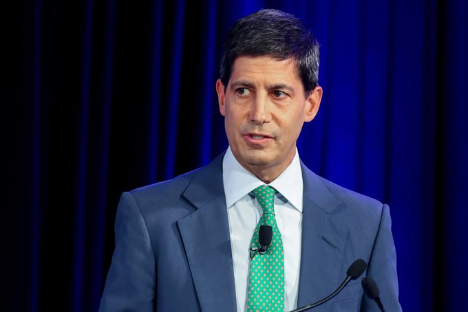 A professional photograph of a man in a blue suit and green polka dot tie speaking at a podium with a microphone, set against a dark blue curtain backdrop.