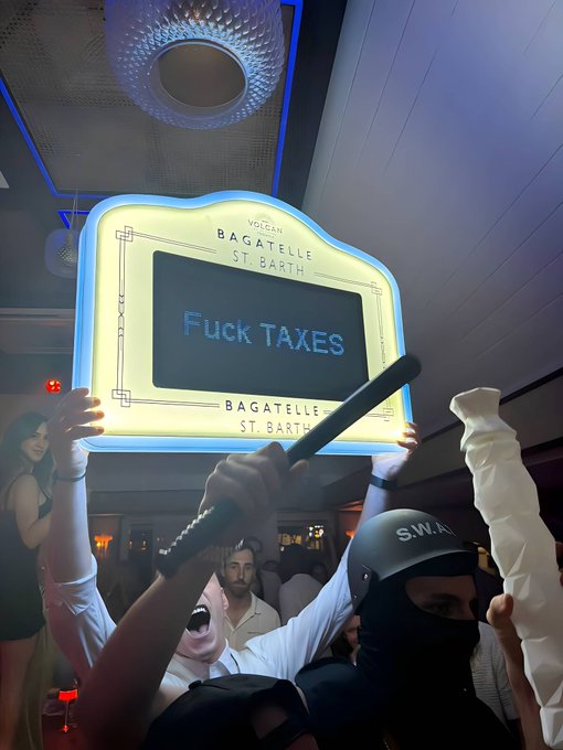 A scene inside Bagatelle St. Barth nightclub where someone holds up an illuminated sign reading 'Fuck TAXES' while a staff member in a SWAT helmet stands nearby.