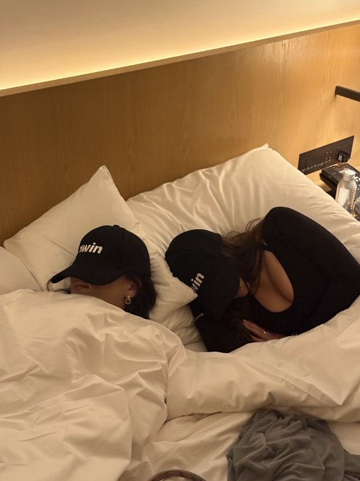 Two people are sleeping side by side under white blankets in a hotel-style bed with a wooden headboard, both wearing black 'Bvlgrin' baseball caps pulled down over their eyes.