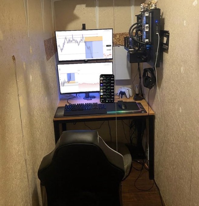 A small home gaming setup in a narrow space features dual monitors displaying charts, a keyboard, mouse, and gaming PC mounted on the wall.