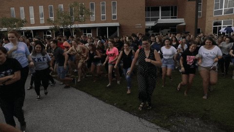 The image shows a large group of people running down a street or sidewalk. The crowd consists of both men and women wearing various colored clothing. Some individuals are carrying backpacks as they run through the scene, while others have umbrellas with them. The overall atmosphere appears to be energetic and lively, possibly due to an event or gathering taking place in the area.