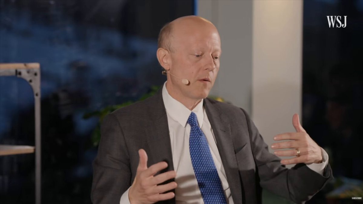 The image shows a man wearing a suit, tie, and earrings giving a speech or presentation. The man appears to be bald with a serious expression as he speaks. The background features a window that allows natural light into the room, creating an atmosphere of professionalism and focus.