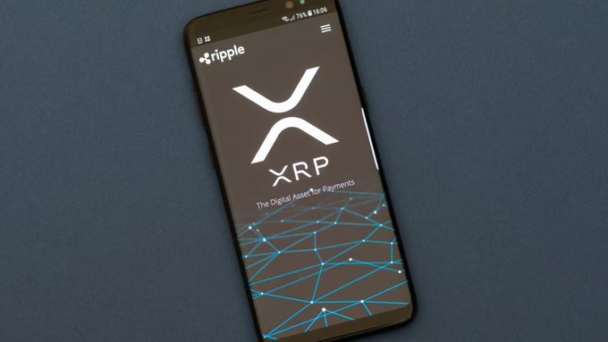 Image of a smartphone displaying the Ripple XRP app on its screen, showing the XRP logo and tagline 'The Digital Asset for Payments' against a network connection graphic, resting on a dark surface.