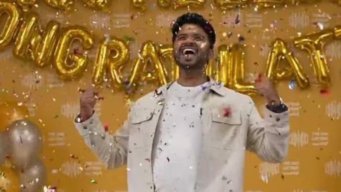 The image features a man standing in front of a gold banner that says "congrats" with confetti falling around him. The man appears to be celebrating an achievement or milestone, as he has his arms raised high in the air. The surrounding environment is colorful and vibrant, with various objects such as balloons and confetti scattered throughout the scene.