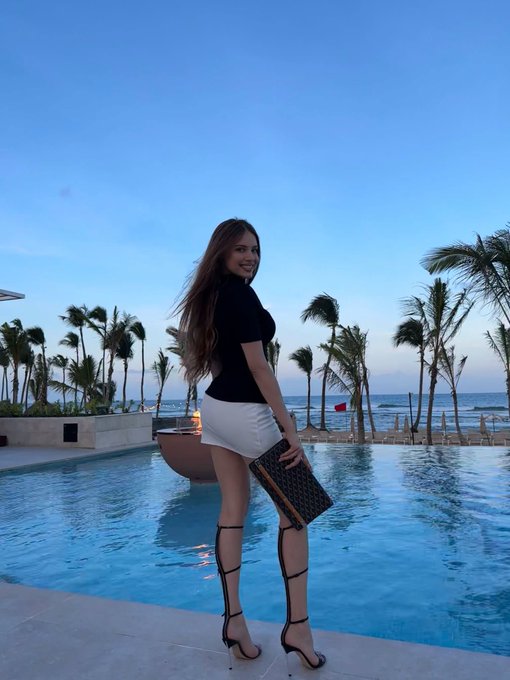 A woman in a black top and white mini skirt stands by a pool with palm trees and the ocean in the background, holding a clutch bag with a monogram pattern.