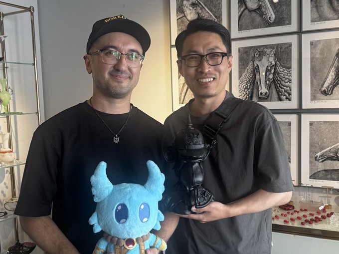 Two men smile together indoors, with the man on the left wearing a "nori" cap and holding a blue horned plush toy, while the man on the right holds a black armored bust, framed by horse artwork in the background—capturing a warm reunion that echoes the post's message of "APE Together Strong" through their friendly bond and shared creative spirit.