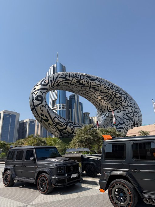 Two dark-colored Mercedes-Benz G-Wagons are parked in front of Dubai's Museum of the Future, a large ring-shaped structure adorned with Arabic calligraphy.