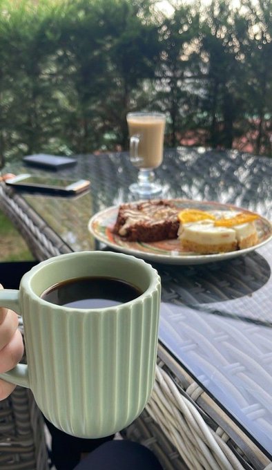 A hand holding a green ribbed mug of black coffee on a glass table, with a plate of food (toast, eggs, and meat) and a glass of latte in the background.