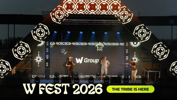 The image captures W Group's "W Fest 2026" stage event with the tagline "The Tribe Is Here," featuring three performers under an illuminated geometric backdrop displaying "W Group" — visually embodying the company's global team gathering described in the post, where diverse members united as one tribe.