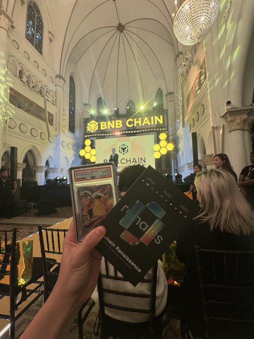 A photograph taken inside a grand hall at a BNB Chain event, showing a person holding a trading card and a 'Chain Renaissance' booklet with the 'fnds' logo.