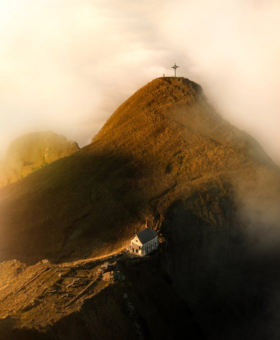 A misty, golden-lit mountain peak topped with a small cross, with a small building situated on a ridge below.