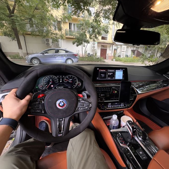 The interior of a high-performance BMW, likely an M5, featuring a black and tan leather cabin with an M-branded steering wheel and infotainment screen, as the driver greets the day with a "gM" from behind the wheel.