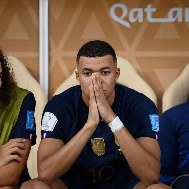 A young soccer player wearing a France national team jersey with the FIFA World Cup Qatar 2022 badge and the number 10 sits on the bench with his hands clasped over his face in a prayerful gesture, with "Qatar" signage visible behind him.