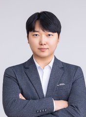 Professional headshot of an Asian man with short black hair, wearing a dark gray blazer over a white collared shirt, arms crossed against a plain light background.