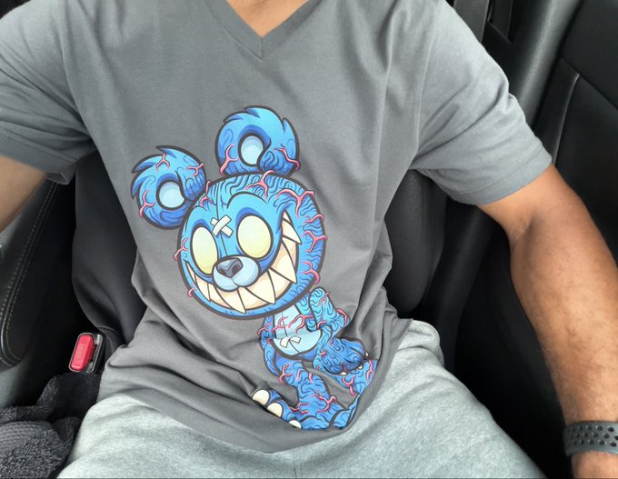 A person wearing a gray v-neck t-shirt featuring a stylized blue cartoon character design with red vein-like patterns, sitting in a black leather seat.