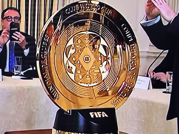 A large golden FIFA Club World Cup trophy on display, with people in suits visible in the background, one taking a photo with a smartphone.