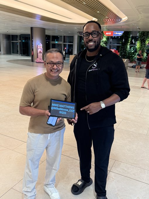 Two men smiling in a mall setting, with one holding a digital sign reading 'MIKE MATTHEWS @MikeMoves_COP $Q3' while standing next to a much taller man in black.