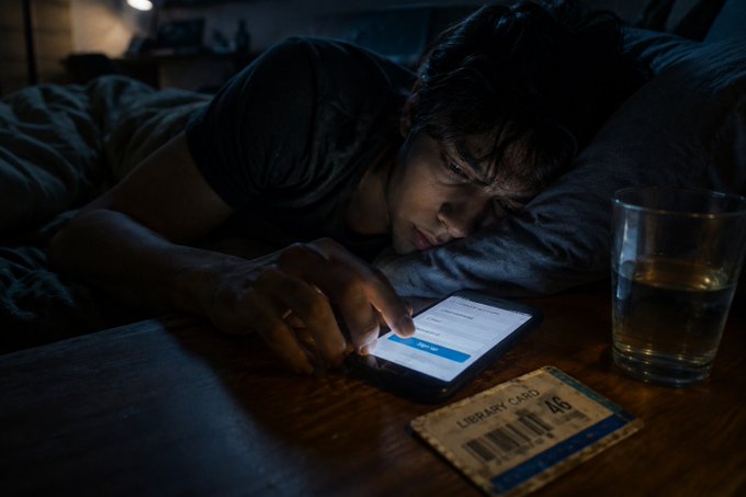 A man lies in bed in a dark room, staring at his smartphone with a tired expression while a glass of amber liquid and a library card rest on the nightstand beside him.