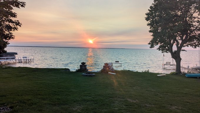 A sunset view over a lake with two people relaxing in lawn chairs on a grassy shore, framed by trees.