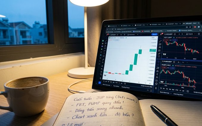 The image shows a dimly lit desk setup at dusk with a laptop displaying TradingView charts for JUP/USDT, FET/USDT, and PUMP/USDT—highlighting a bullish green candle on JUP contrasting with bearish red candles on FET and PUMP. Beside the laptop sits a coffee mug, a pen, and an open notebook with handwritten Vietnamese notes referencing JUP, PUMP, FET, and DeFi tokens, visually reinforcing the post's message about how quickly capital flow direction can shift across crypto tokens over the weekend.