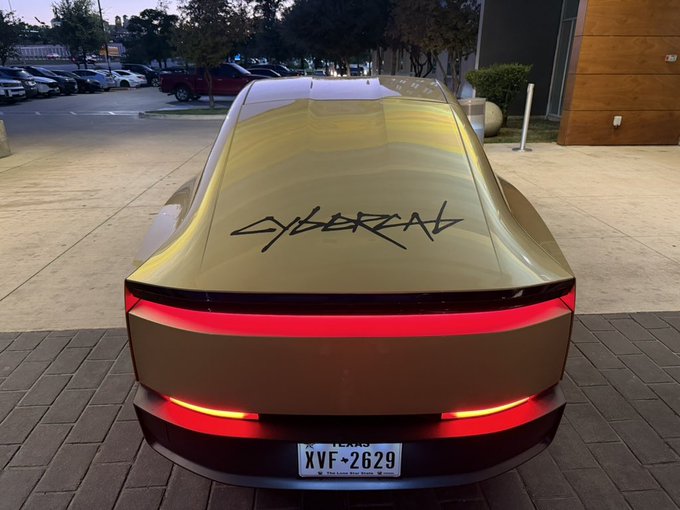 The rear view of a gold/champagne-colored Tesla Cybertruck with "CYBERTRUCK" graffiti-style text on its tailgate and red LED light bar illuminated, displaying a Texas license plate "XVF-2629".