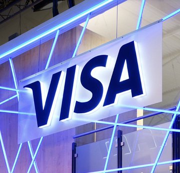An illuminated blue and white 'VISA' logo sign is mounted on a wall with blue neon light accents, likely at a trade show or corporate event.