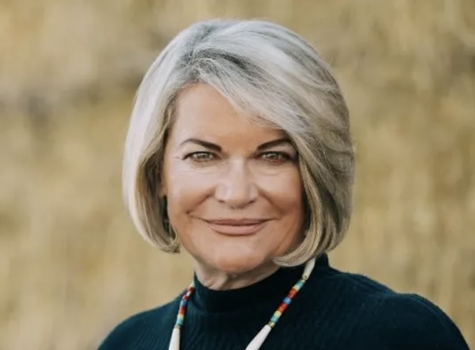 A smiling woman with shoulder-length gray hair, wearing a black turtleneck and colorful beaded necklace, poses for a portrait against a textured beige background. The image accompanies a post about Senator Lummis expressing her support for advancing the CLARITY Act.