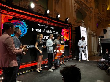 The image shows a group of people standing around a stage with a large banner that says "Proof of Pitch 2016". The event appears to be a pitch competition, as there are several individuals holding microphones and presenting their ideas. One person on the right side of the photo is wearing glasses and has a microphone in front of them. A man stands next to this individual, possibly part of the judging panel or an audience member. Another man can be seen standing further back from the stage. The group appears to be engaged in a discussion about the event or the competition itself.