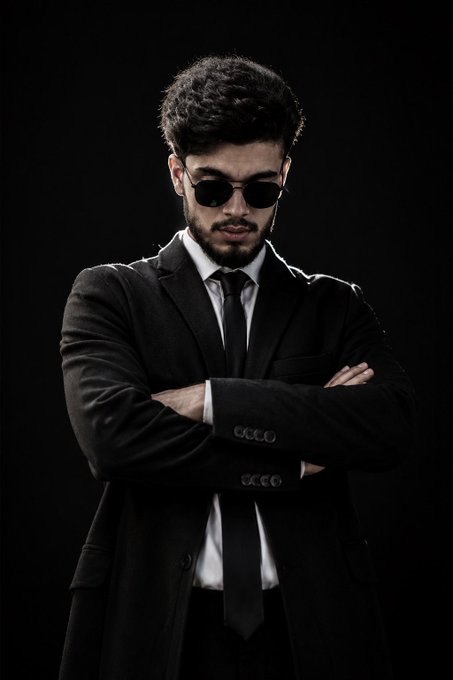 A young man wearing a black suit, white shirt, black tie, and dark sunglasses stands with his arms crossed against a black background.