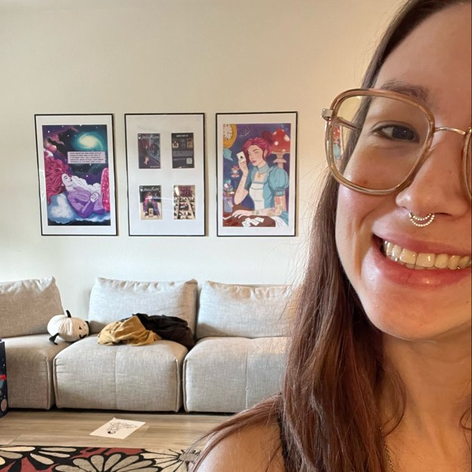 Close-up selfie of a smiling woman wearing glasses and a nose ring, with a gray couch and three framed illustrations on the wall behind her.