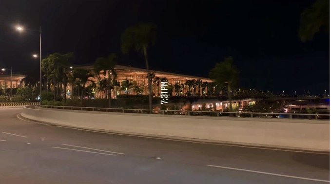 Nighttime photo of what appears to be an airport terminal with palm trees, viewed from a curved roadway, with a timestamp overlay reading '7:31PM'.