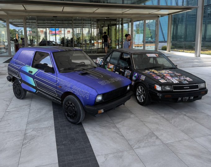 Two customized cars—a blue "SOLANA"-branded hatchback and a black vehicle covered in stickers with the license plate "DK 1739 NW" and "DODGE" badge—are parked side by side on a paved plaza in front of a modern glass building. The image ties to the post's $TCAR campaign by showcasing the "Solana" themed trenchcar alongside a sticker-covered ride, visually representing the "ride or take the bus" choice between custom crypto-themed vehicles and public transit.