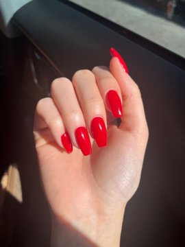 Close-up photograph of a hand displaying long, glossy red almond-shaped nails, lit by sunlight against a dark background.