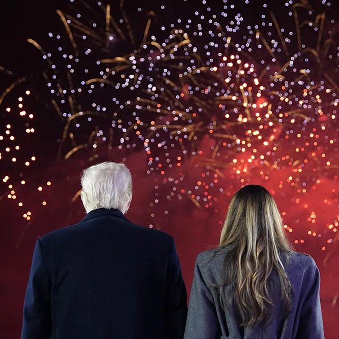 Two people viewed from behind, watching a colorful fireworks display explode against a dark red sky.