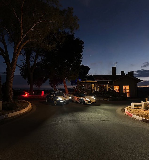 Two sports cars driving along a curved road at dusk with their headlights on, passing a lit house under a deep blue sky.