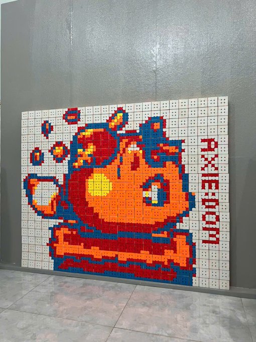A pixel art mosaic of the Bitcoin logo (orange and yellow 'B' symbol) created on what appears to be physical Rubik's cubes or similar cubelets, mounted on a gray wall.
