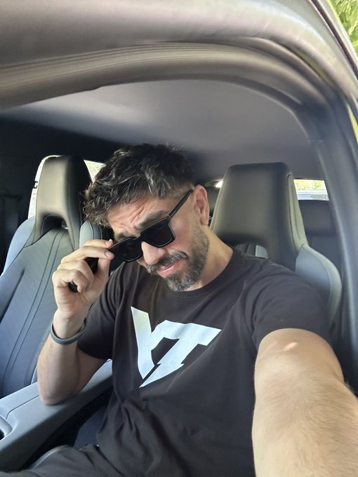 A selfie of a bearded man in sunglasses and a black 'W' logo t-shirt sitting in the driver's seat of a car.